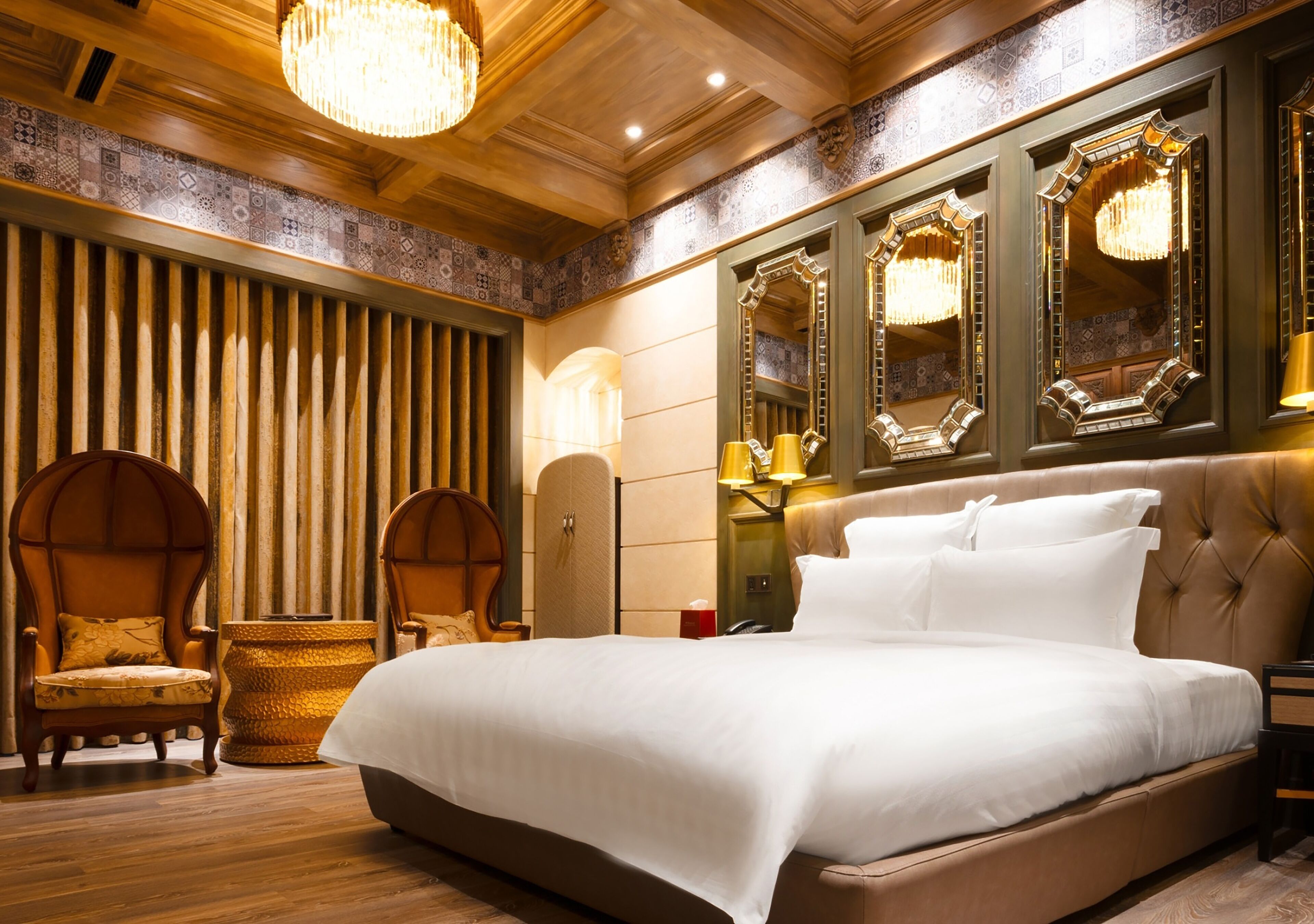 A large bed with white linens, two upholstered chairs, a chandelier, and ornate mirrors.