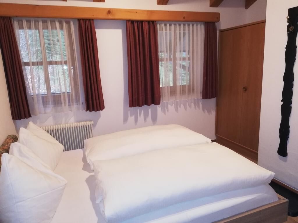 A bedroom with a wooden headboard, a bed with white linens, two windows with sheer and dark curtains, and a radiator.