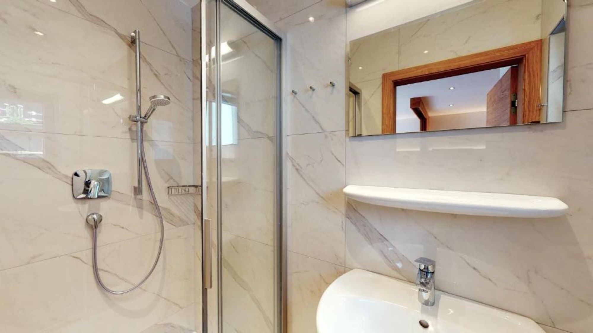 A modern bathroom with a glass-enclosed shower, a marble-tiled wall, a wooden-framed mirror, and a white pedestal sink.