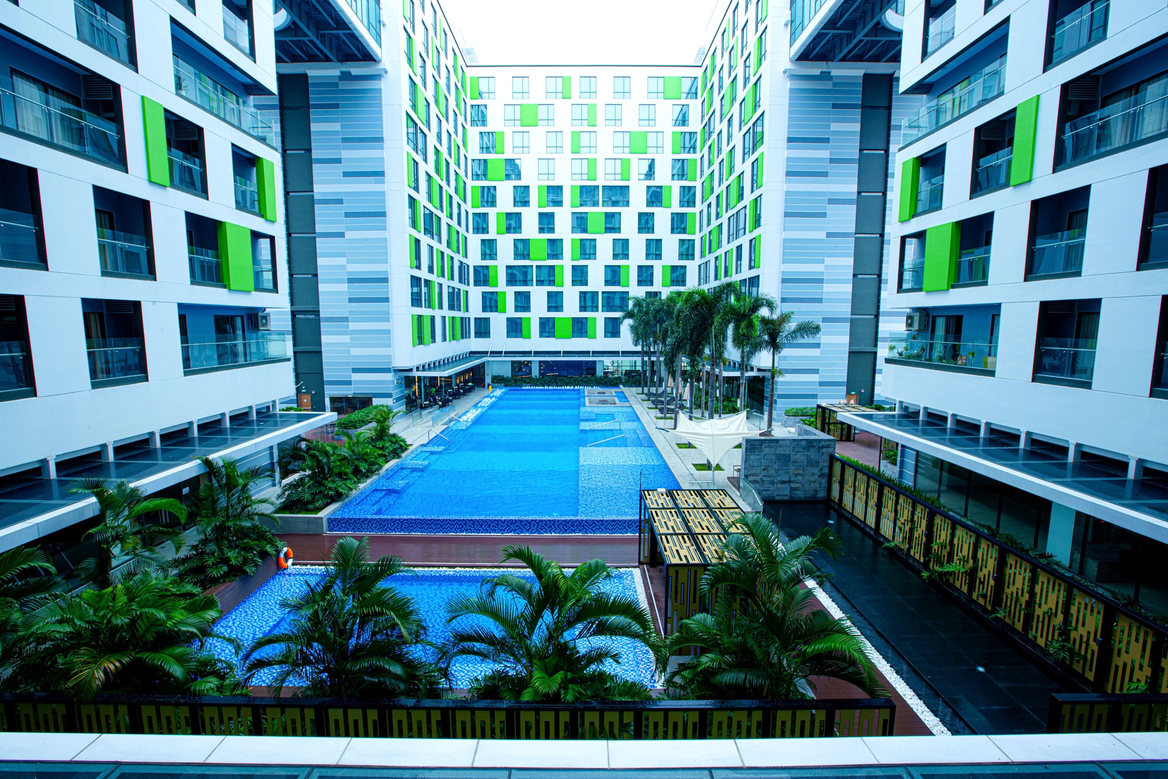 A modern hotel with a central courtyard featuring a swimming pool, surrounded by lush greenery and palm trees.