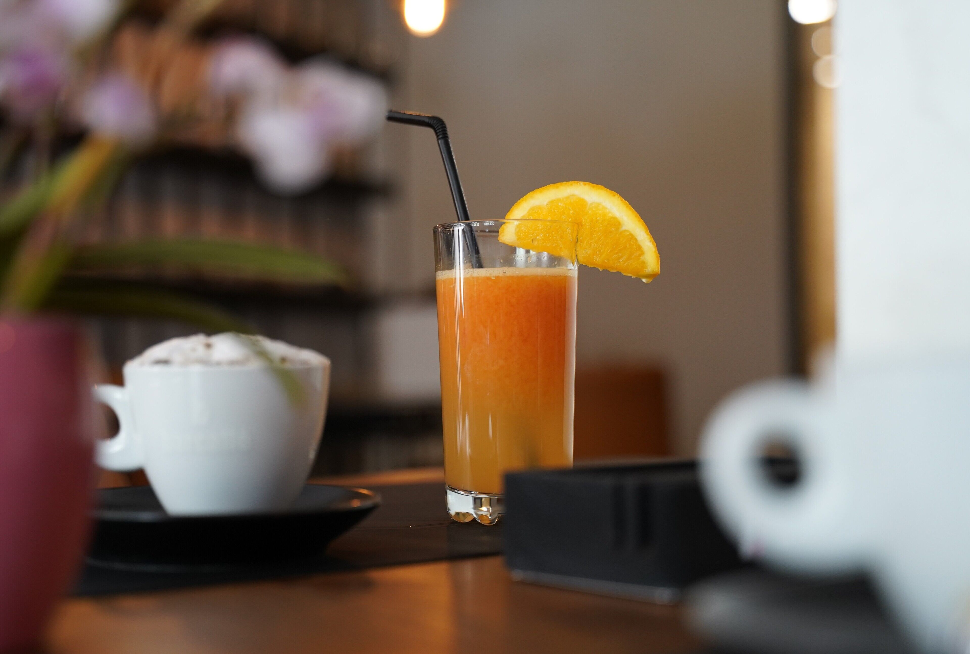 A glass of orange juice with a straw and a slice of orange on the rim, a white cup, and a small black object on a table.