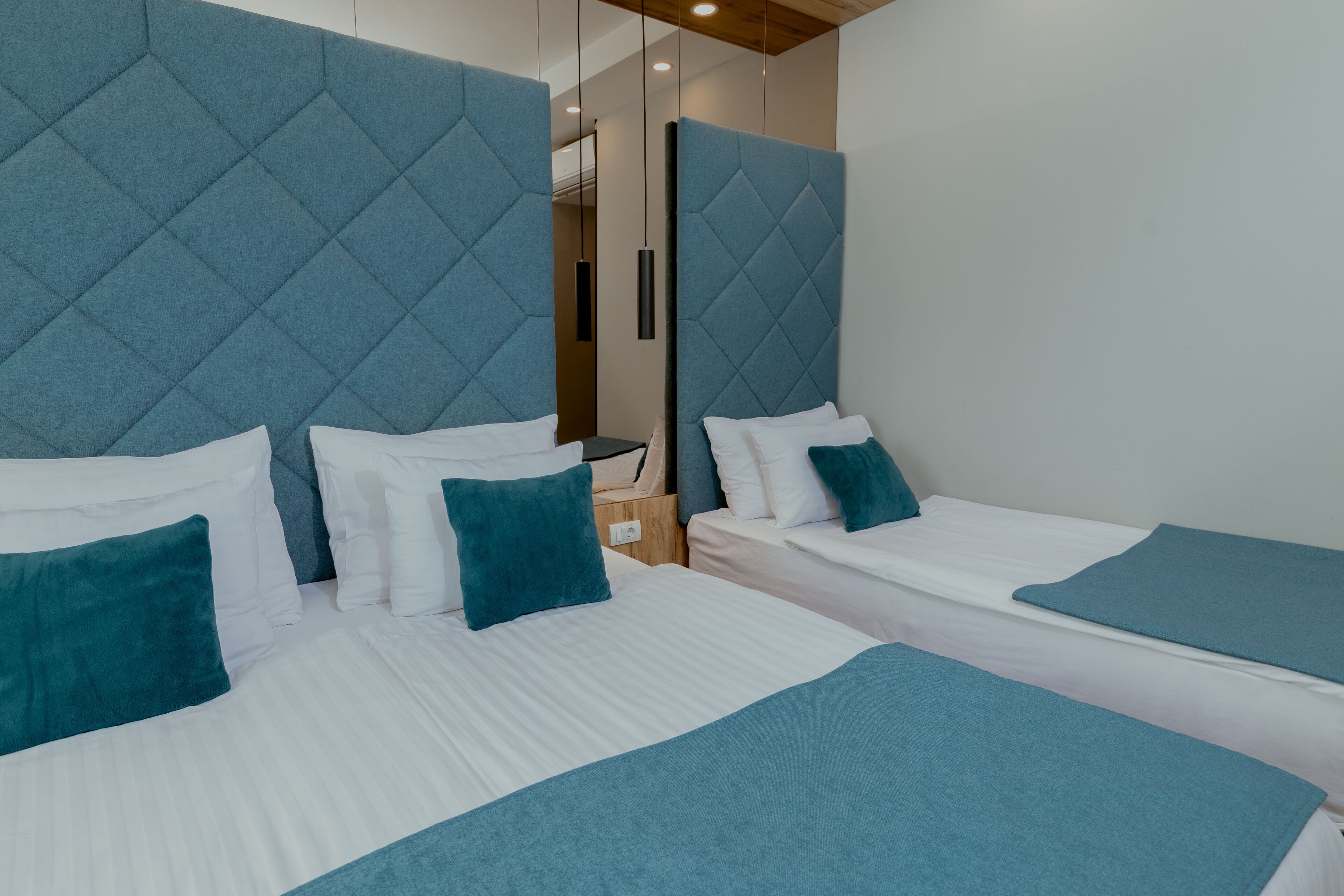 Quarto de hotel com duas camas, cada uma com roupa de cama branca e detalhes verde-água. O quarto apresenta um design moderno, com uma cabeceira grande e acolchoada e teto de madeira.