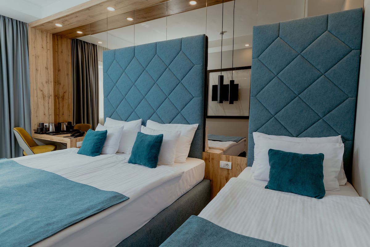 A hotel room with two beds, each with a blue headboard, white bedding, and teal throw pillows. There is a wooden desk with a chair, a small table, and a wall-mounted TV.