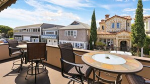 A rooftop patio with wicker chairs and a glass-top table, overlooking a row of buildings with a mix of architectural styles. - Air conditioned Balboa Island Flat, Steps to Balboa Island Ferry, w/Parking. Amazing Location! (Newport Beach)