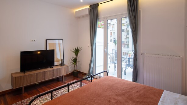 A modern bedroom with a large bed, a TV on a wooden console, a potted plant, and a balcony with a view of buildings. - Phaedrus Living: City Luxury Flat Mavromichali (Athens)