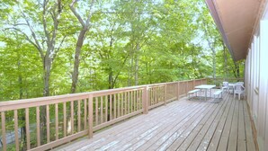 Une terrasse en bois avec une rambarde, des tables et des chaises blanches, et une vue sur une zone boisée.