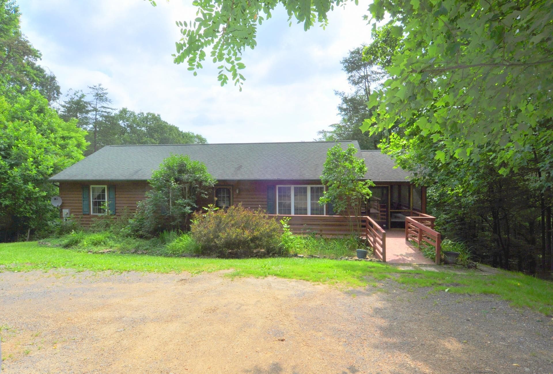 A wooden cabin with a porch and a gravel driveway, surrounded by trees and greenery.