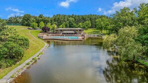 A serene lakeside setting with a pool and a building surrounded by lush greenery. - Country Comfort -country Escape! (Hedgesville)