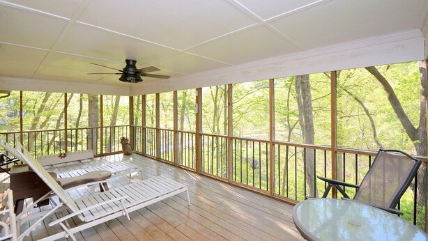 A covered porch with a ceiling fan, wooden railings, and a view of trees.