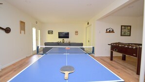 A ping pong table with a paddle in the foreground, a sofa in the background, and a foosball table to the right.