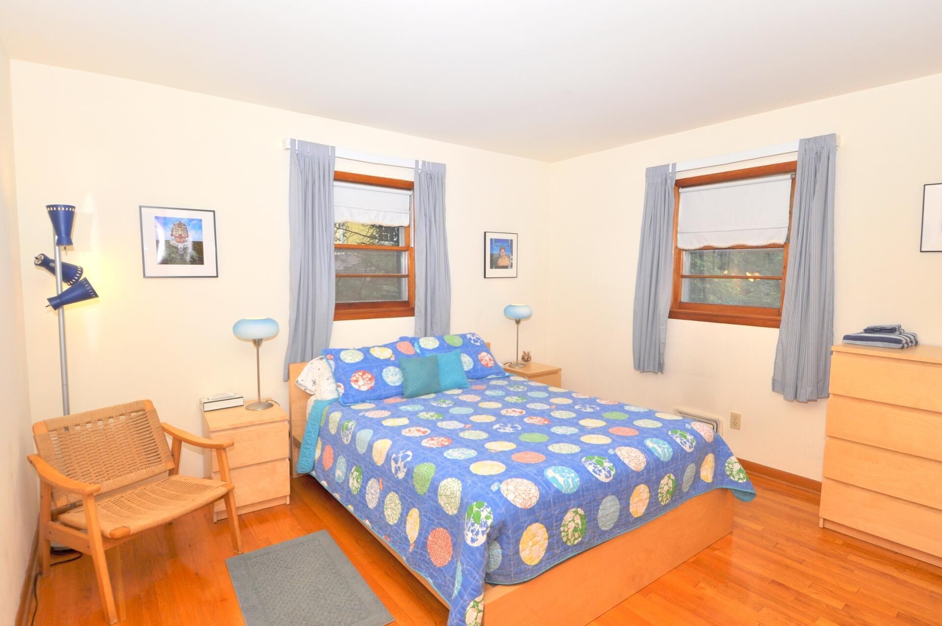 A bedroom with a bed, two bedside tables, a chair, a dresser, and two windows with curtains.