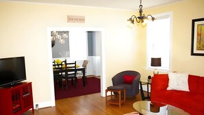 A living room with a red sofa, a glass coffee table, a TV on a red cabinet, a dining area with a table and chairs, and a hallway with a sign. - Aunt Beas Bungalow - Right in Town! (Berkeley Springs)