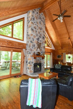 A cozy cabin interior with a stone fireplace, wooden ceiling, and large windows.