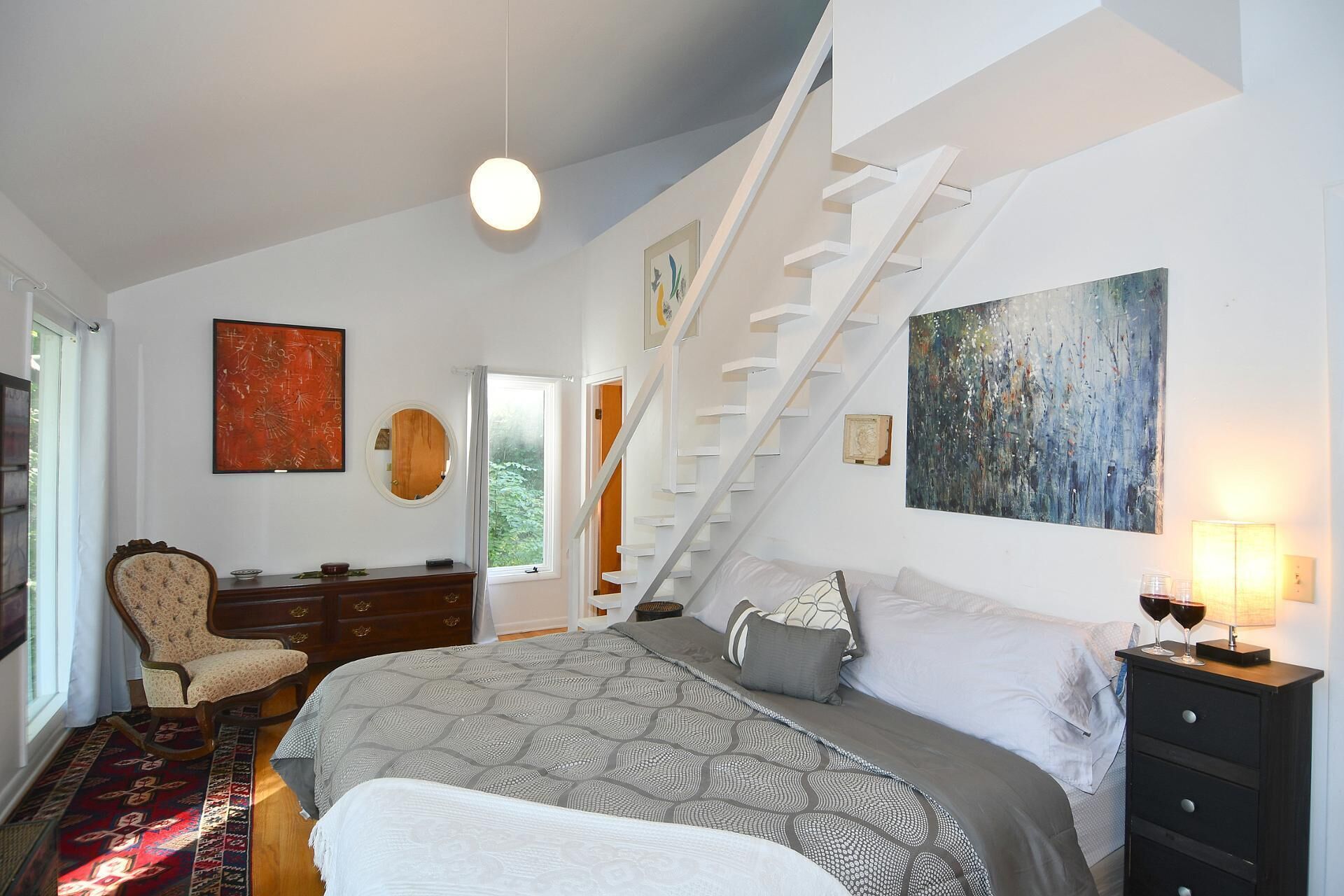 A bedroom with a bed, a chair, a dresser, a staircase, and artwork on the walls.