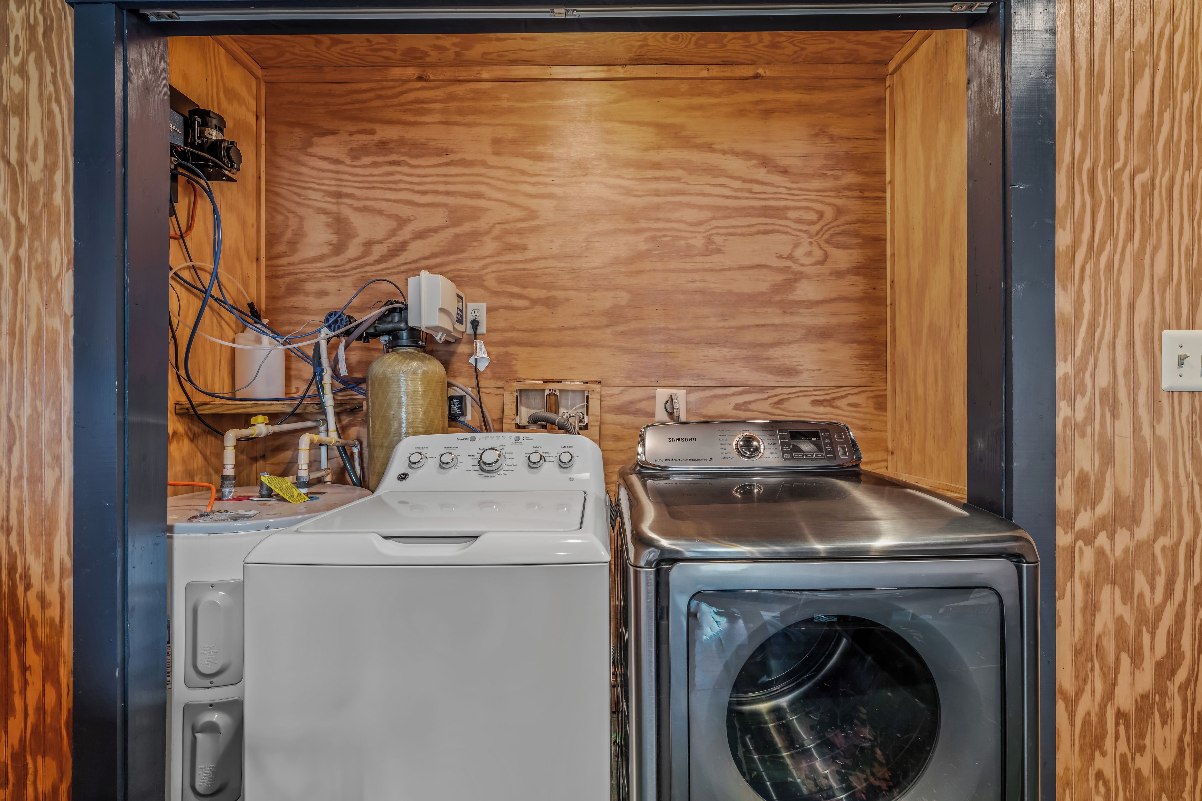 A compact laundry room with a washing machine and a dryer, wooden walls, and visible plumbing.