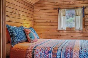 A wooden cabin bedroom with a bed, pillows, and a window with curtains. - Whispering Hills - Couples Getaway (Berkeley Springs)