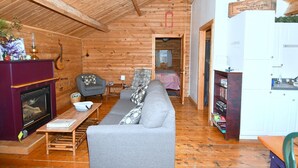 A cozy log cabin interior with a fireplace, a sofa, a dining table, and a kitchen area. - Whispering Hills - Couples Getaway (Berkeley Springs)