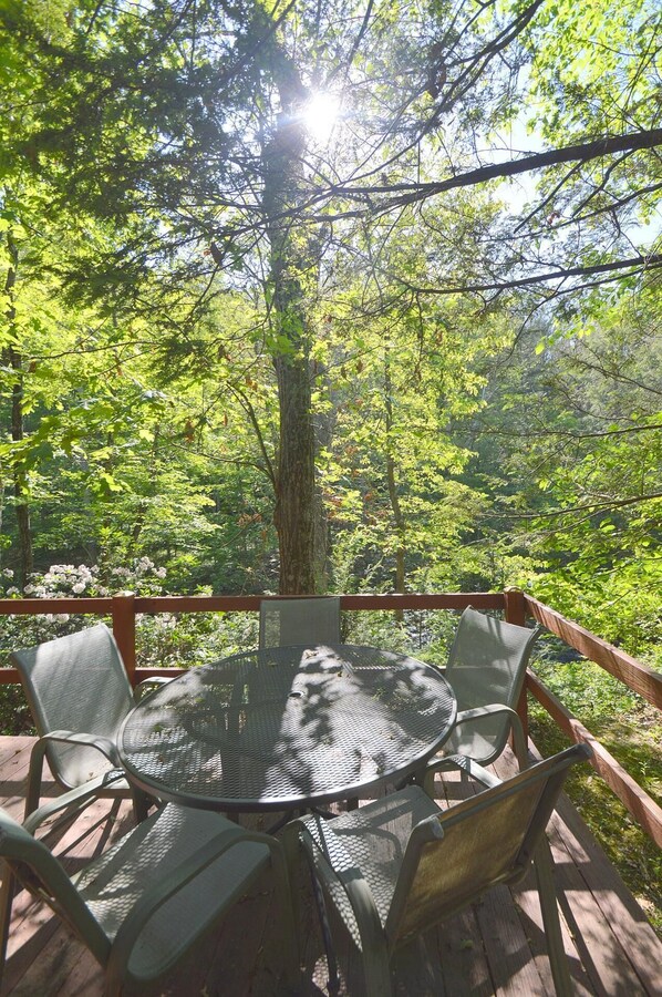 Une table ronde en métal, entourée de six chaises, est posée sur une terrasse en bois, elle-même bordée d’arbres verdoyants et d’une rambarde en bois.
