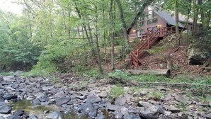 Uma cabana com escadaria de madeira que dá para um deck, rodeada por floresta densa e um riacho rochoso.