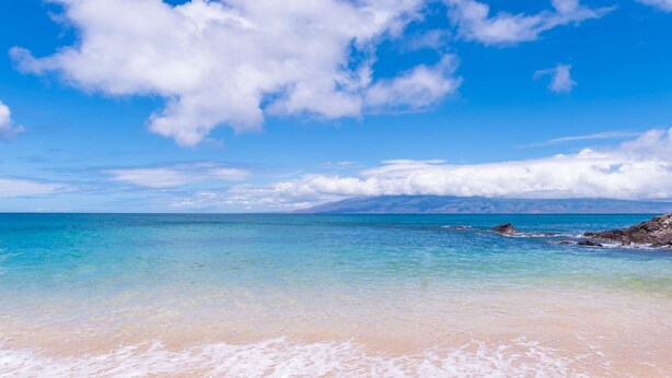 Ein Strand mit klarem türkisblauem Wasser, einem sandigen Ufer und einer Bergkette im Hintergrund.