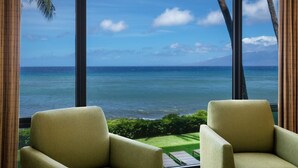 Een kamer met een groot raam met uitzicht op de oceaan en een berg in de verte. Op de voorgrond staan twee groene fauteuils.