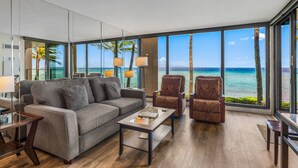 Una sala de estar moderna con un gran ventanal con vista a la playa, un sofá gris, dos sillones estampados y una mesa de centro.