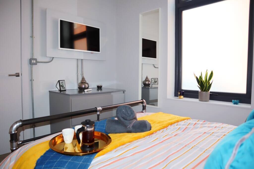 A modern hotel room with a bed, a bedside table, a TV mounted on the wall, and a window with a plant on the sill.