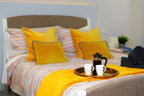 Una cama con ropa de cama a rayas y almohadas amarillas, una bandeja con dos tazas y una cafetera, y una planta en la esquina.
