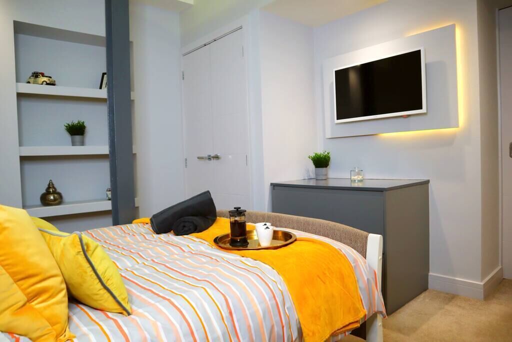 Un dormitorio con una cama, una mesita de noche, un televisor montado en la pared y un estante con objetos decorativos.