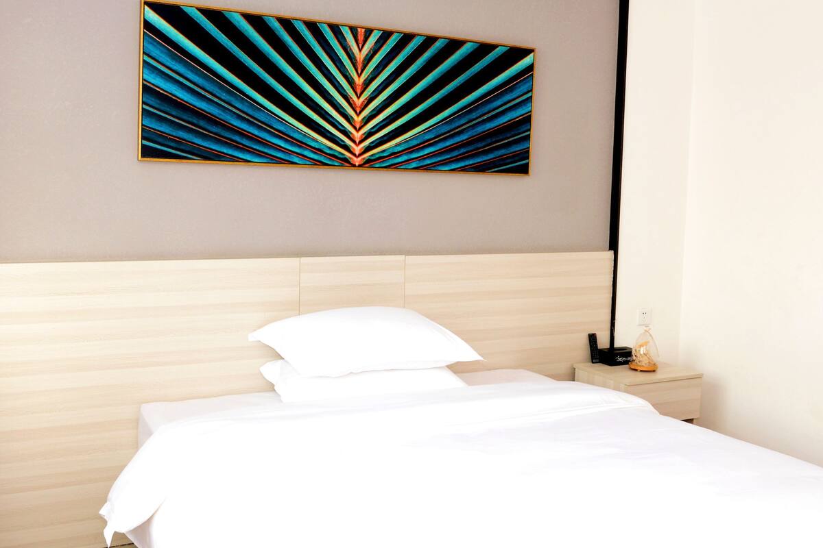 Une chambre d’hôtel moderne avec une tête de lit en bois, une grande peinture abstraite au mur, une table de chevet avec une lampe et un lit parfaitement fait.