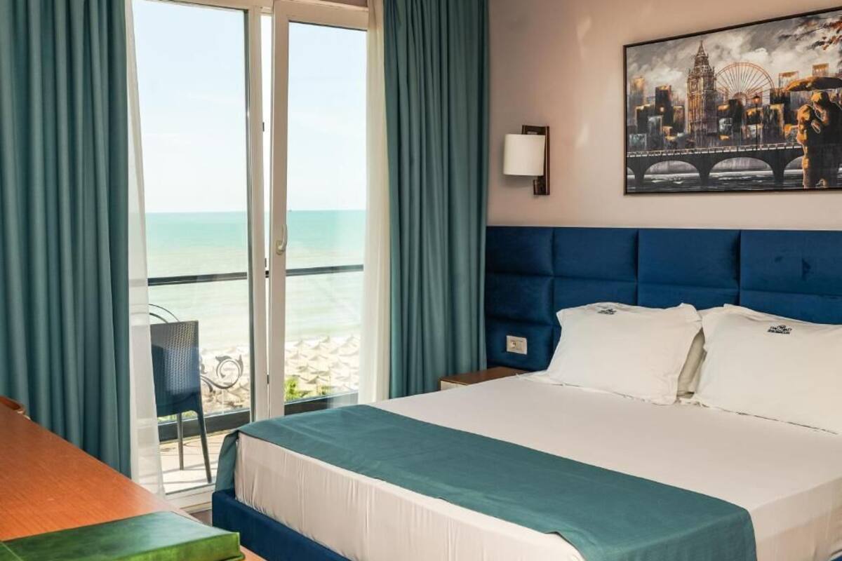A hotel room with a bed, a view of the sea through a sliding door, a cityscape painting on the wall, and a chair by the door.
