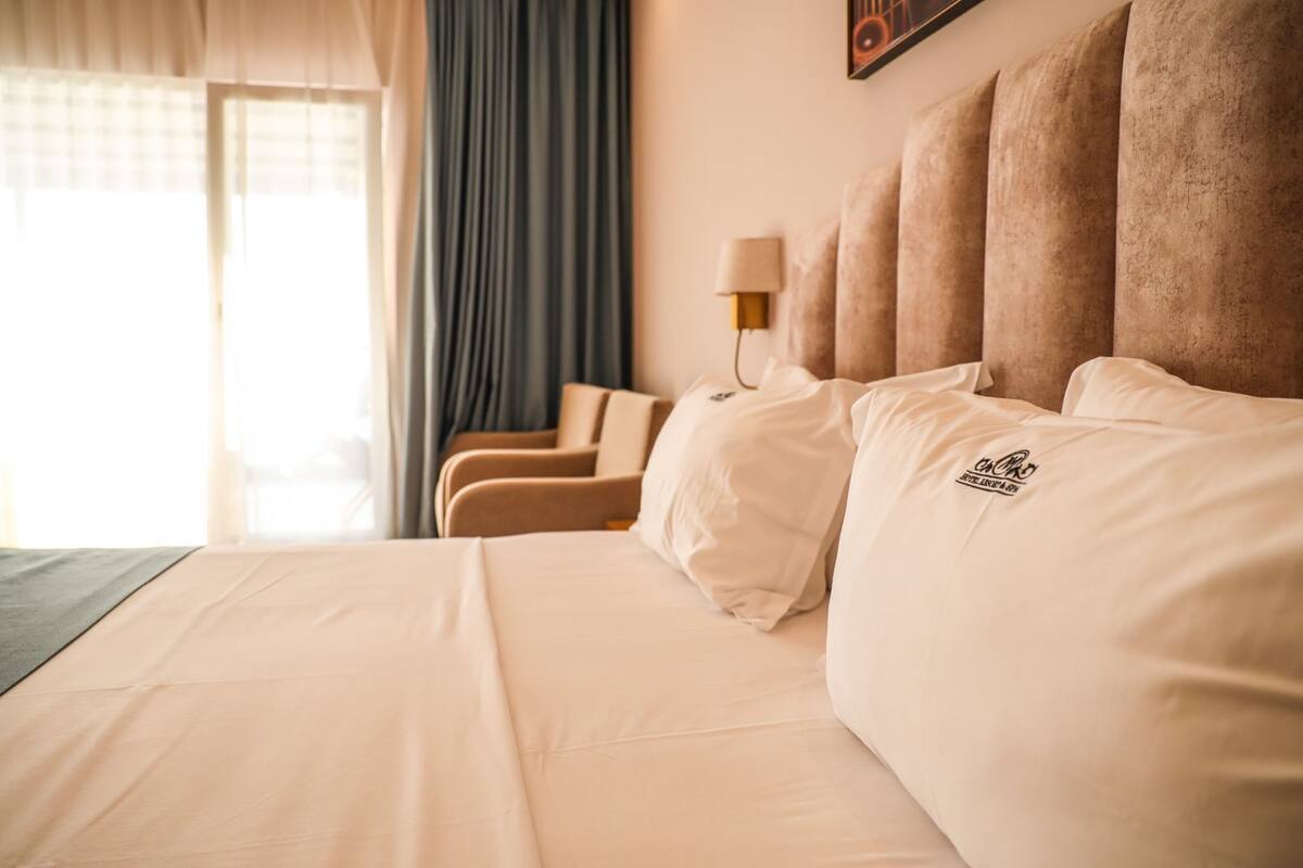 A hotel room with a large bed, multiple pillows, a bedside lamp, and a window with curtains.