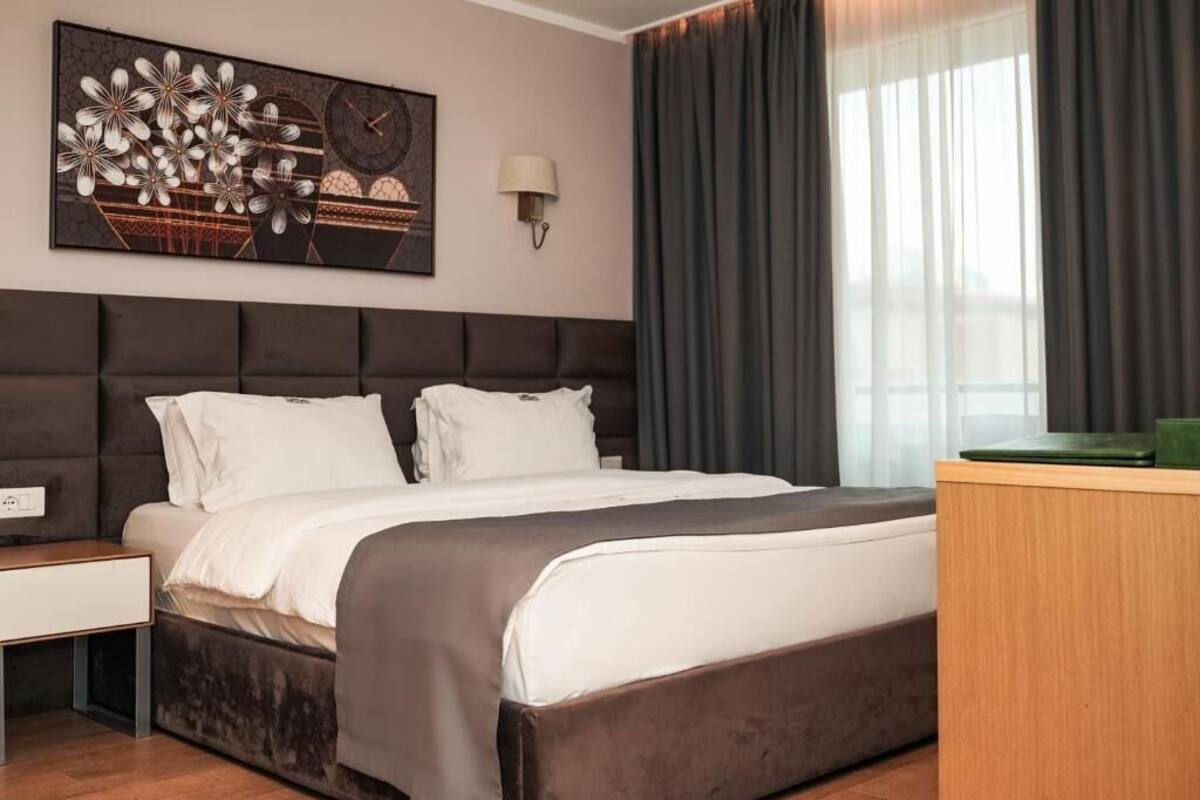A hotel room with a large bed, a nightstand, a wall-mounted lamp, and a window with curtains.