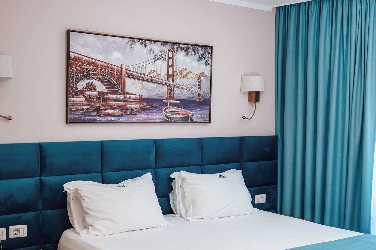 A hotel room with a large bed, a headboard, a nightstand, a lamp, a painting of a bridge, and a window with curtains.