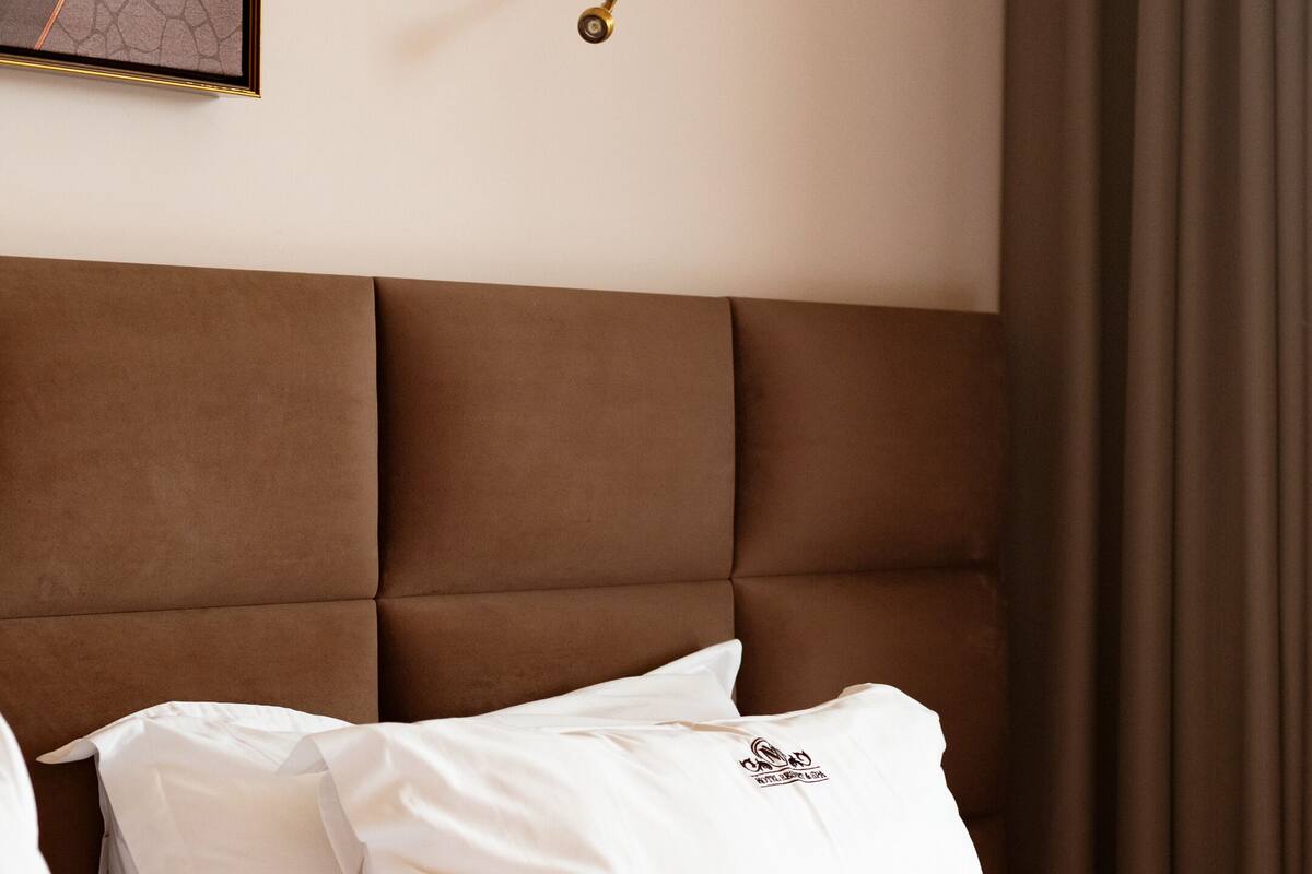 A neatly made bed with white linens and a single decorative pillow, a wall-mounted lamp, and a framed artwork above the bed.