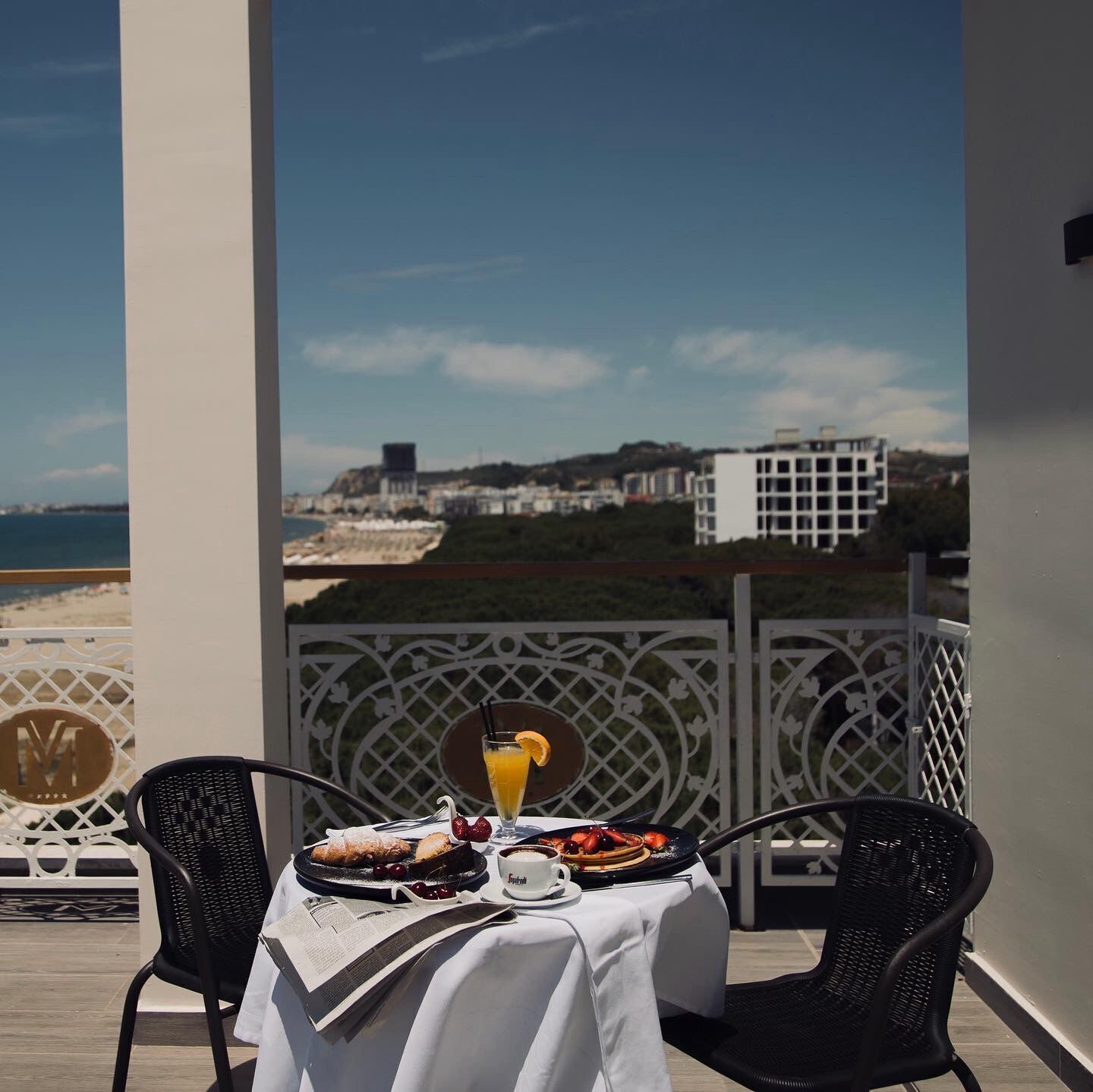 A table set with breakfast items and a drink on a balcony overlooking a beach and buildings.