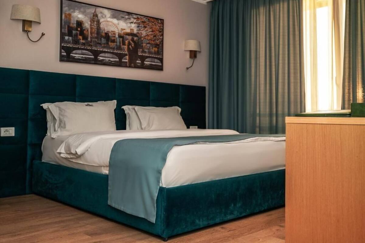 A neatly made bed with a teal headboard and white bedding, a cityscape painting above it, and a wooden nightstand to the right.