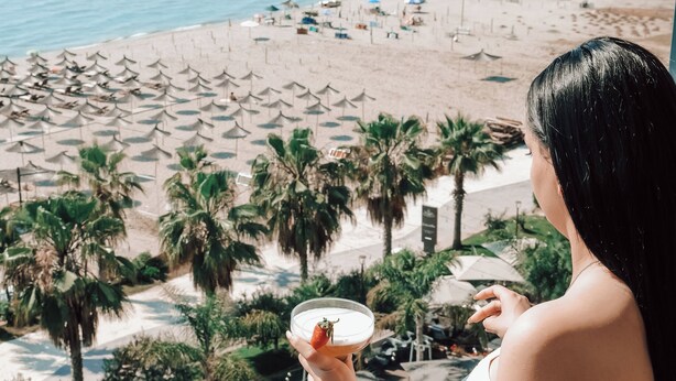 Iemand houdt een drankje vast op een balkon met uitzicht op een strand met parasols en palmbomen.