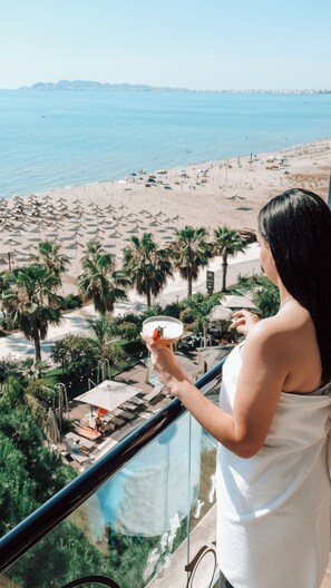 Une personne tenant un verre sur un balcon donnant sur une plage, avec des parasols et des palmiers.