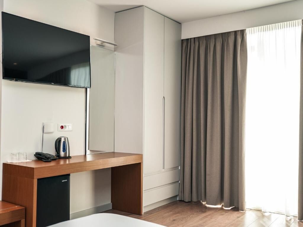 A hotel room with a flat-screen TV, a wooden desk with a kettle, a phone, and a mini-fridge.