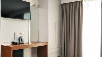 A hotel room with a flat-screen TV, a wooden desk with a kettle, a phone, and a mini-fridge.