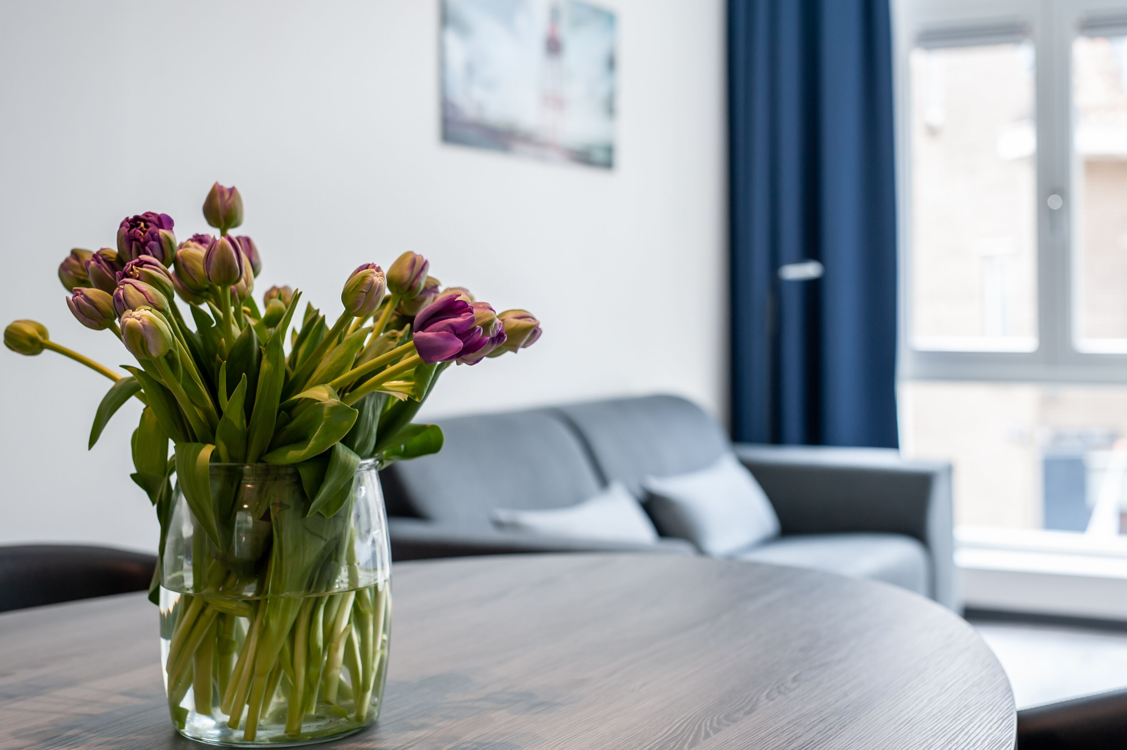 Vista despejada de una habitación con una mesa redonda de madera, un jarrón con tulipanes y un sofá gris al fondo.