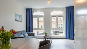 Quarto moderno com uma mesa de jantar, um sofá e uma planta em vaso. Há janelas amplas com cortinas e um quadro na parede.