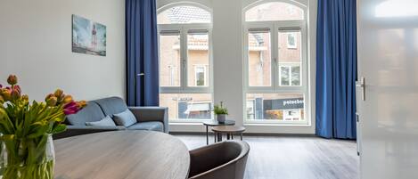 Una habitación moderna con una mesa de comedor, un sofá y una planta en maceta. Cuenta con ventanas amplias con cortinas, y un cuadro en la pared.