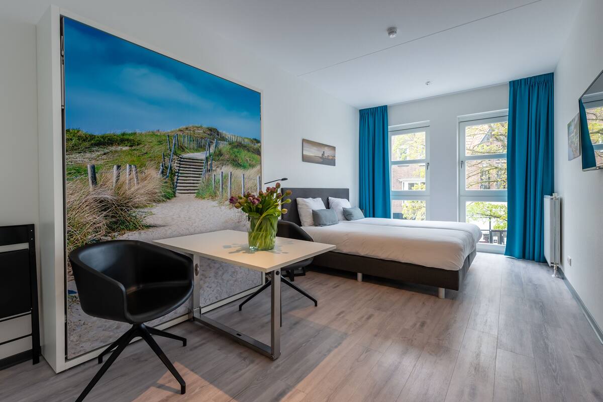 Ein modernes Hotelzimmer mit einem Bett, einem Schreibtisch und einem Sessel. An der Wand hängt ein Gemälde, das eine Strandansicht zeigt.