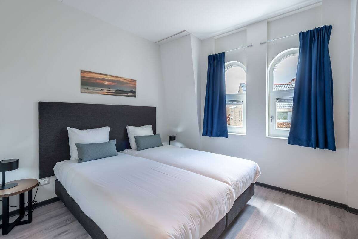 Ein modernes Hotelzimmer mit einem großen Bett, einem Schreibtisch und Blick auf eine Stadtlandschaft durch bogenförmige Fenster.