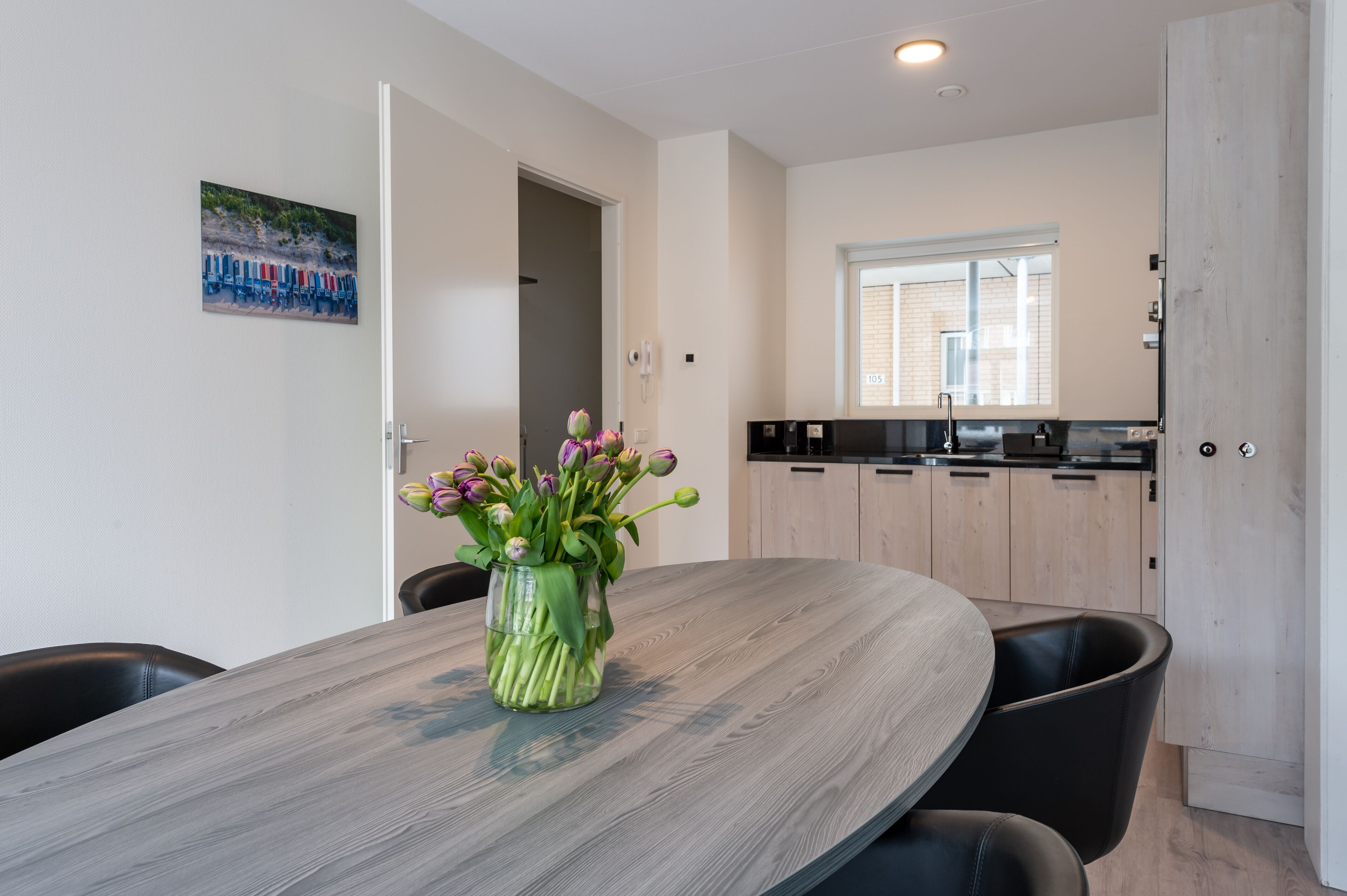 A dining area with a round table, chairs, a vase with flowers, a painting on the wall, and a kitchen in the background.