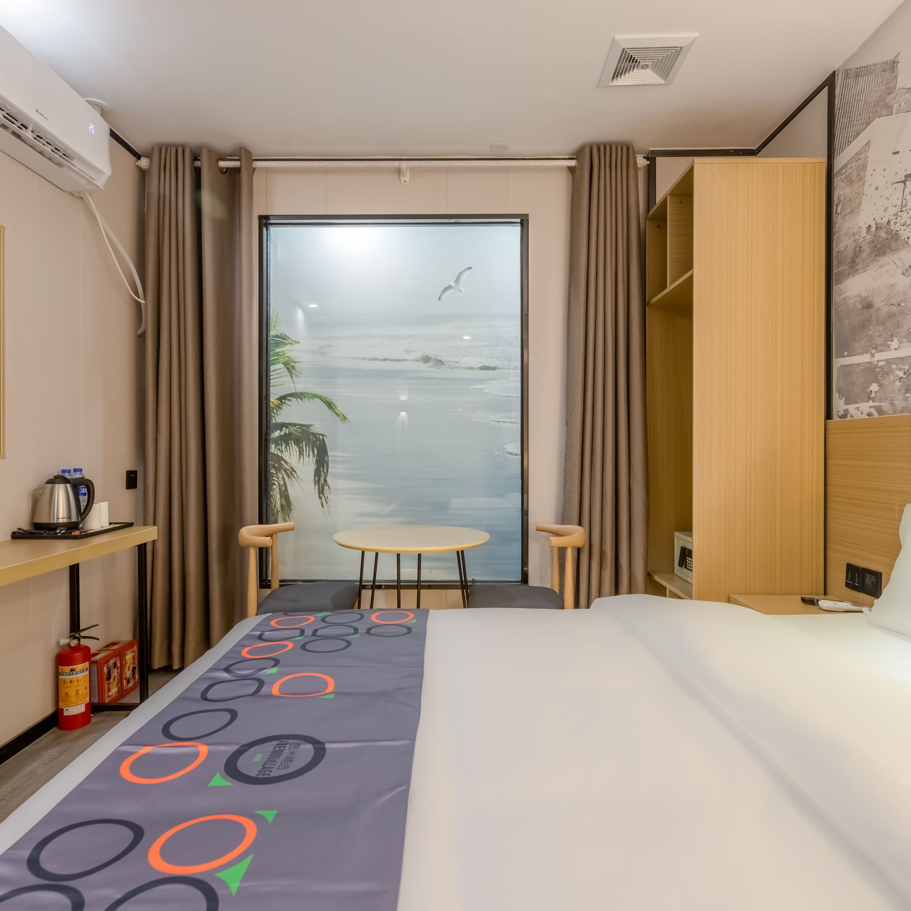 A hotel room with a bed, a small table, two chairs, a view of the beach, and a fire extinguisher.