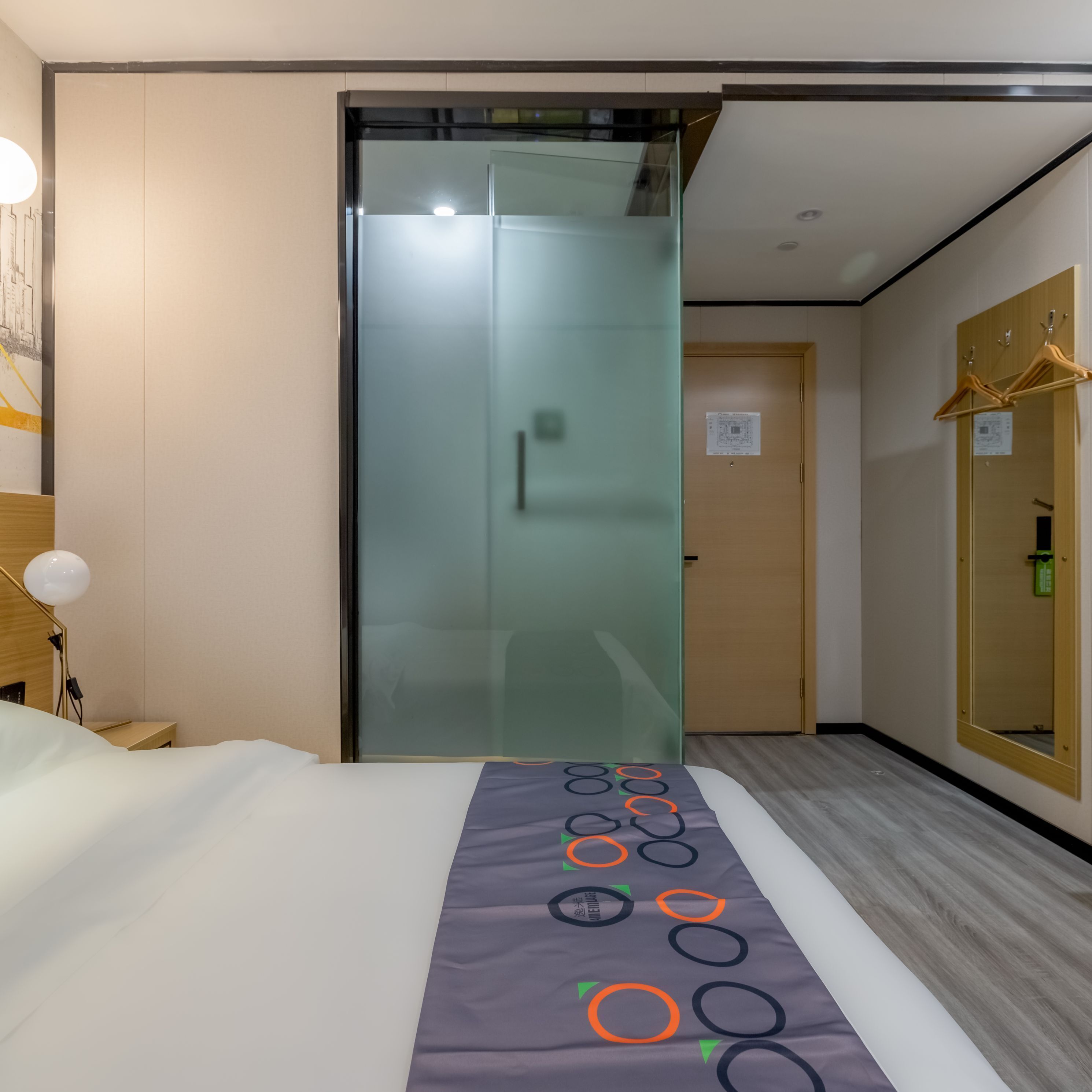 A hotel room with a bed, a desk with a lamp, a sliding glass door, and a wooden wardrobe with a mirror.