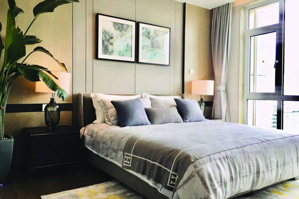 A modern hotel room with a large bed, two framed abstract paintings, a potted plant, and a large window with curtains.